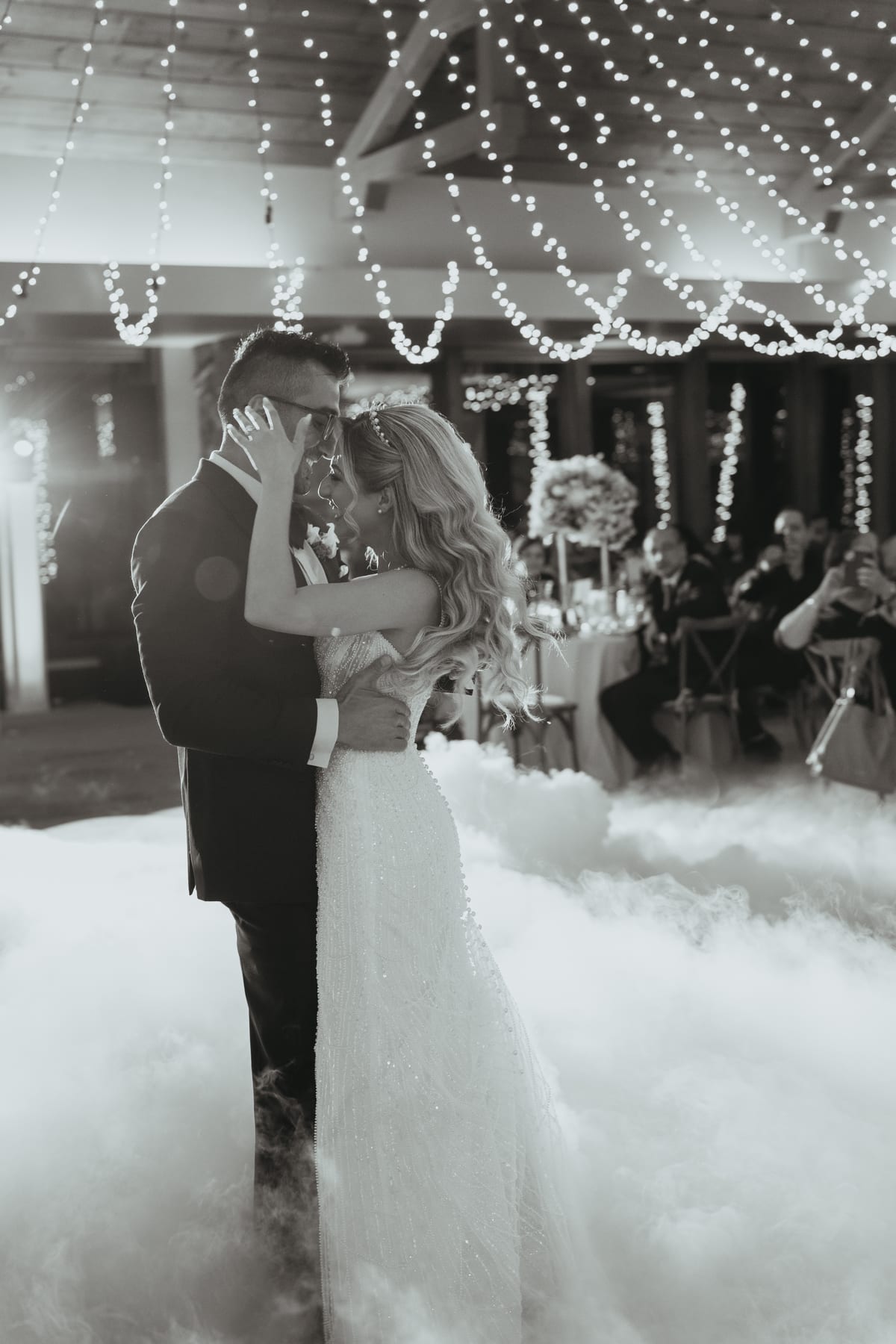 Bride and groom sharing their first dance on a fog-covered floor beneath string lights, black and white