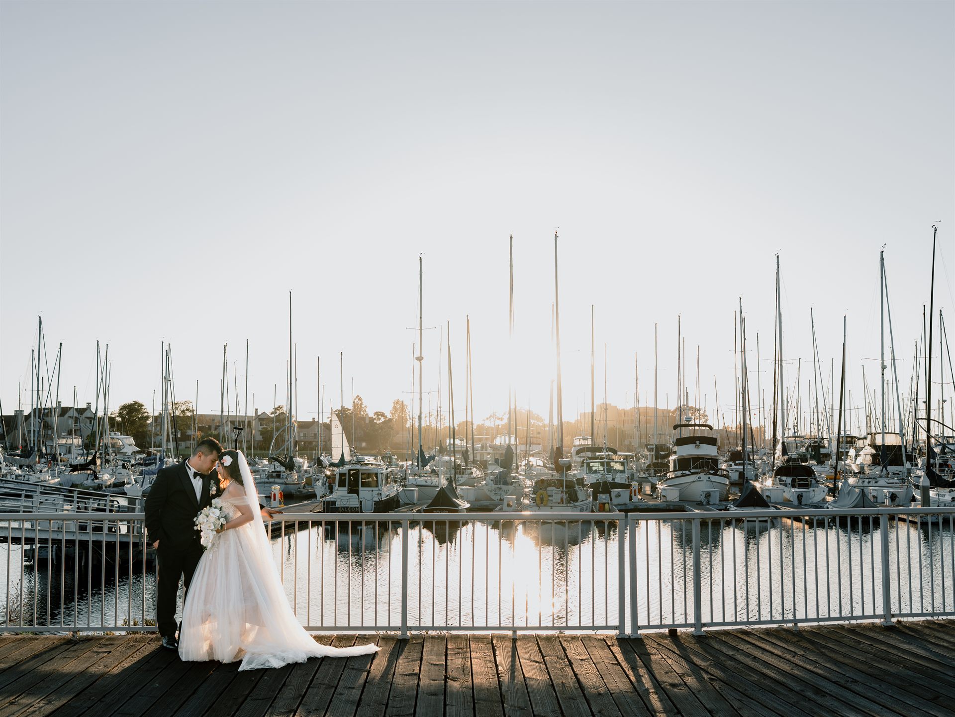 Bride and groom on the marina boardwalk at golden hour, sailboat masts behind them