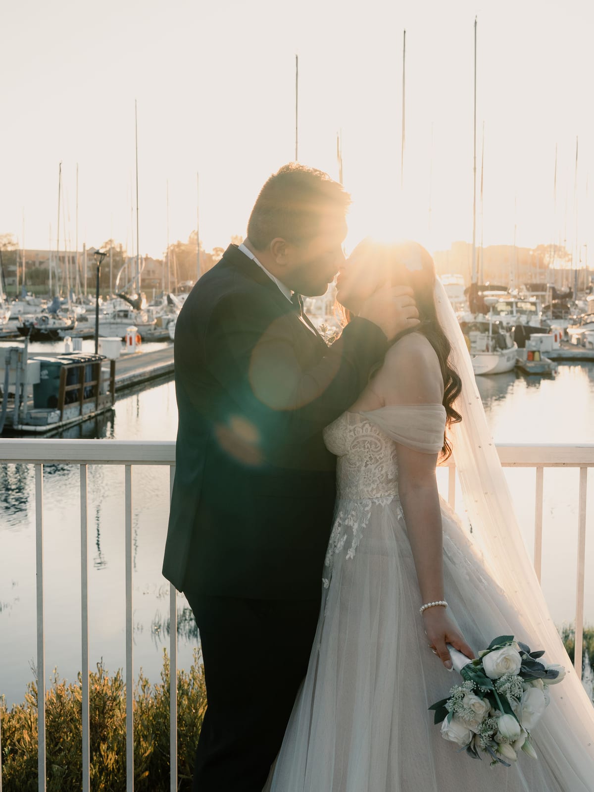 Bride and groom kissing at sunset on the marina railing, sailboat masts glowing behind them