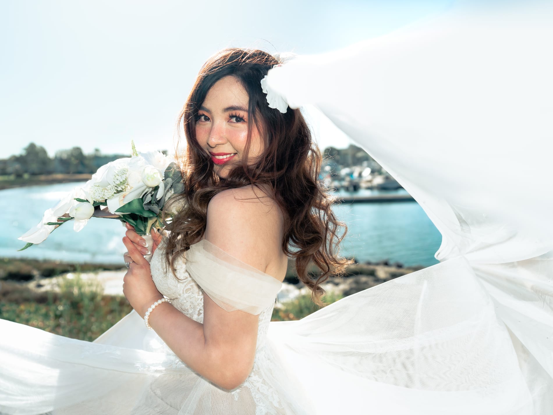 Bride holding her white bouquet as her veil sweeps across the frame, the harbour behind her