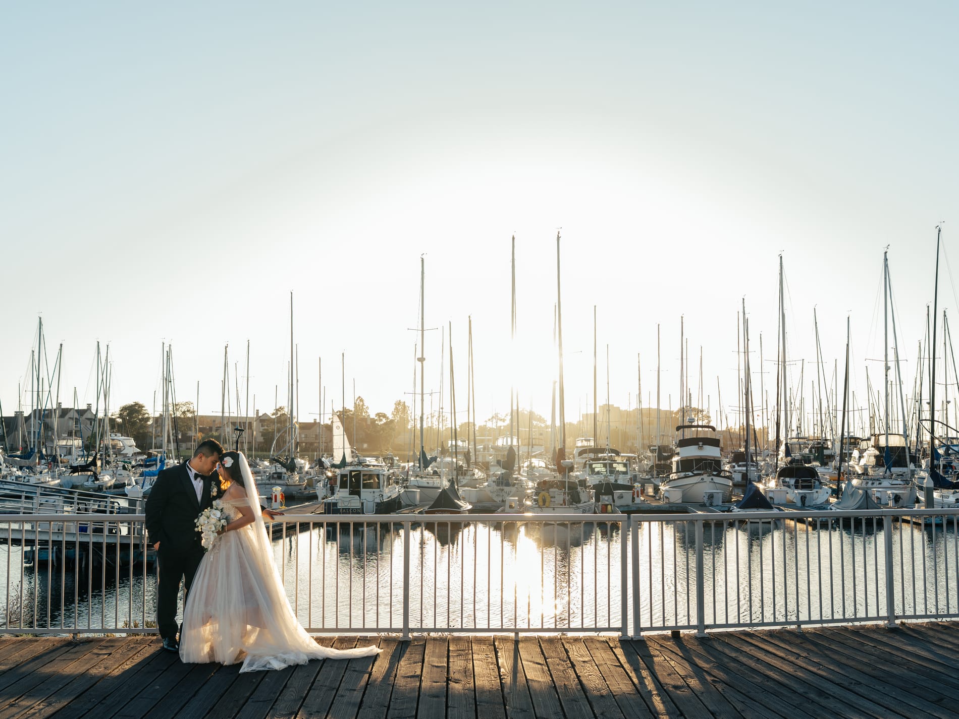 Bride and groom small on the marina boardwalk, sailboat masts and the low sun filling the frame