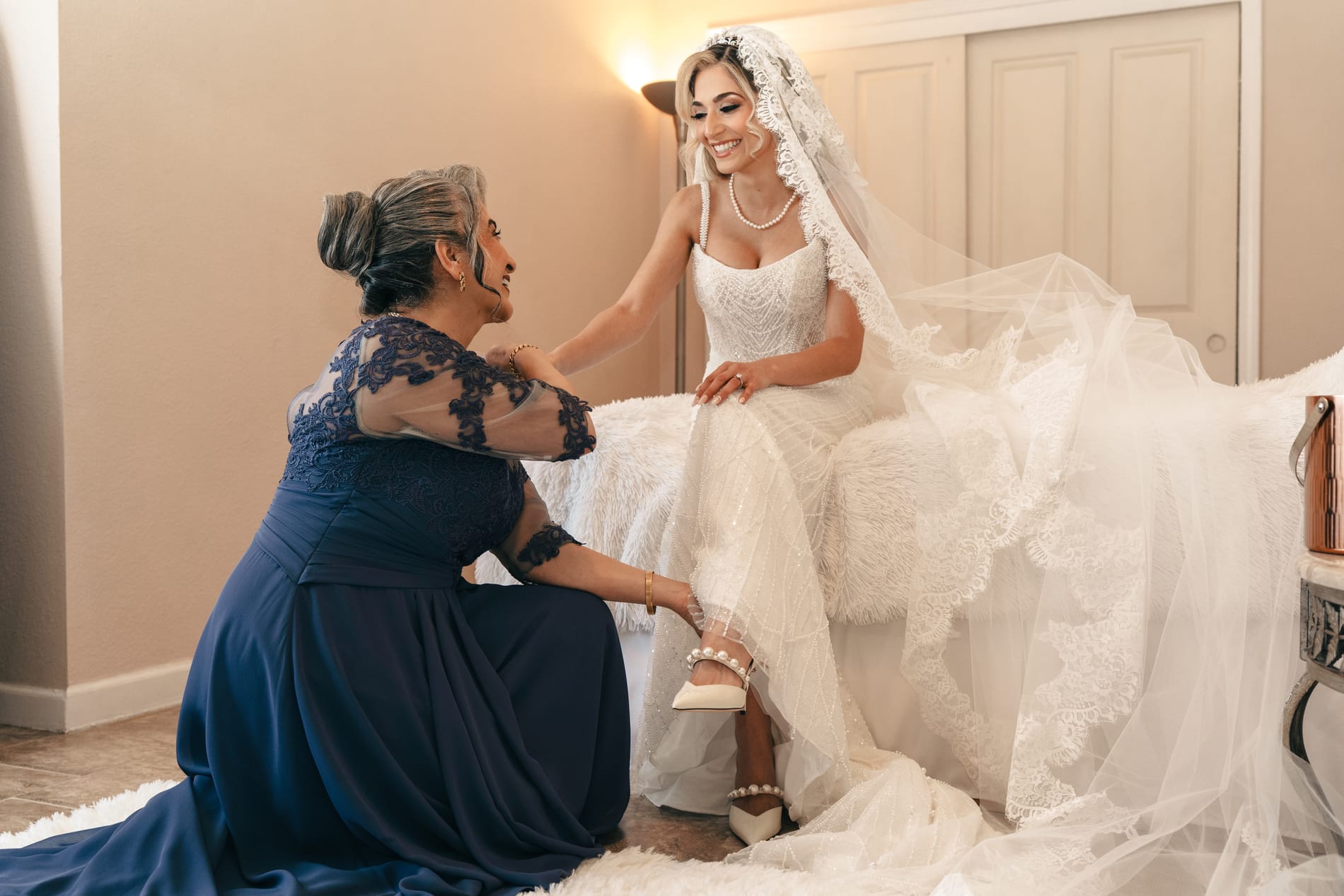 Bride's mother kneeling in navy lace to fasten her shoe while the bride laughs on the bed