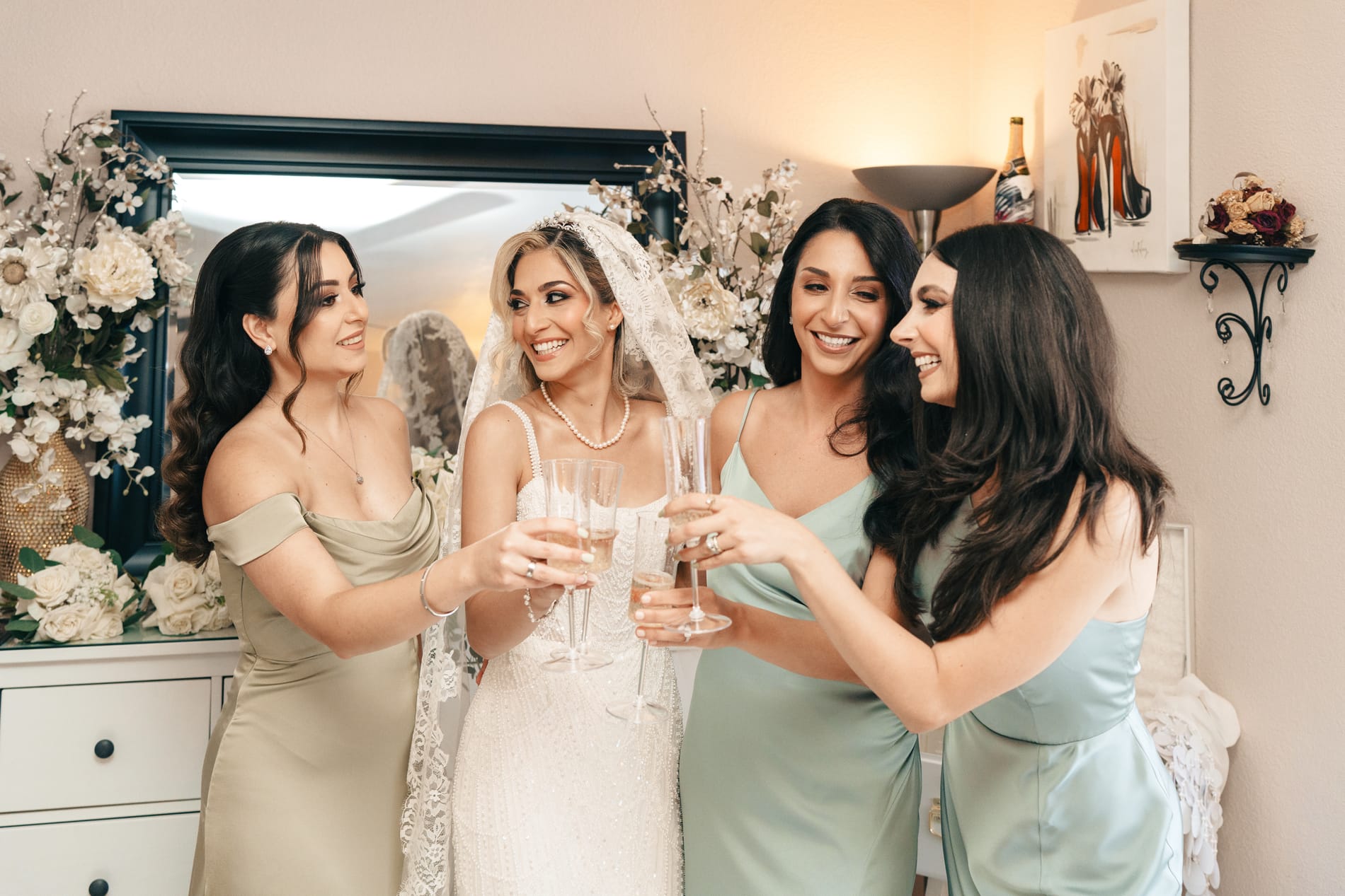 Bride and three bridesmaids in sage dresses laughing over a champagne toast