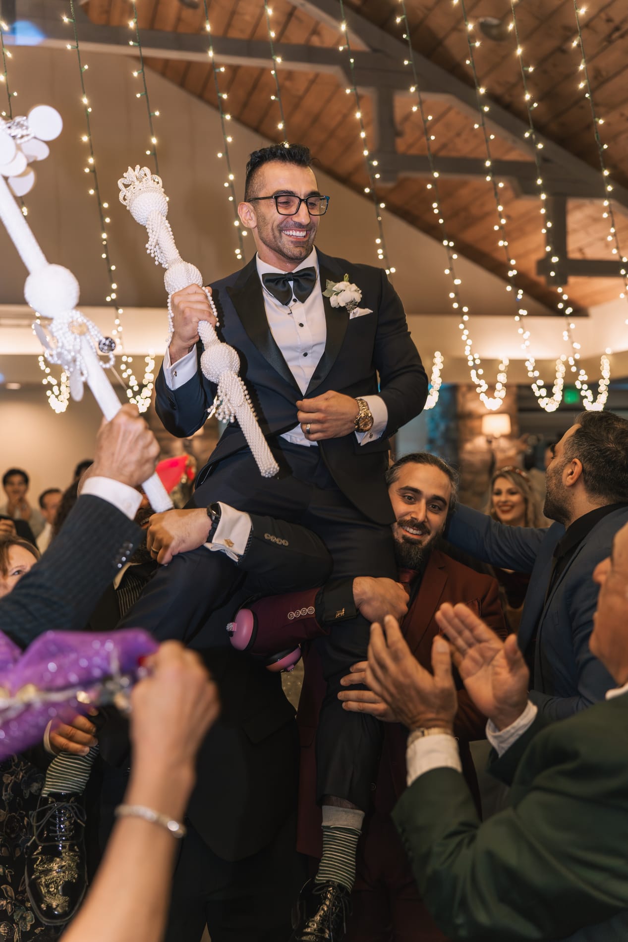 Groom lifted on his friends' shoulders holding the pearl scepter beneath the string lights
