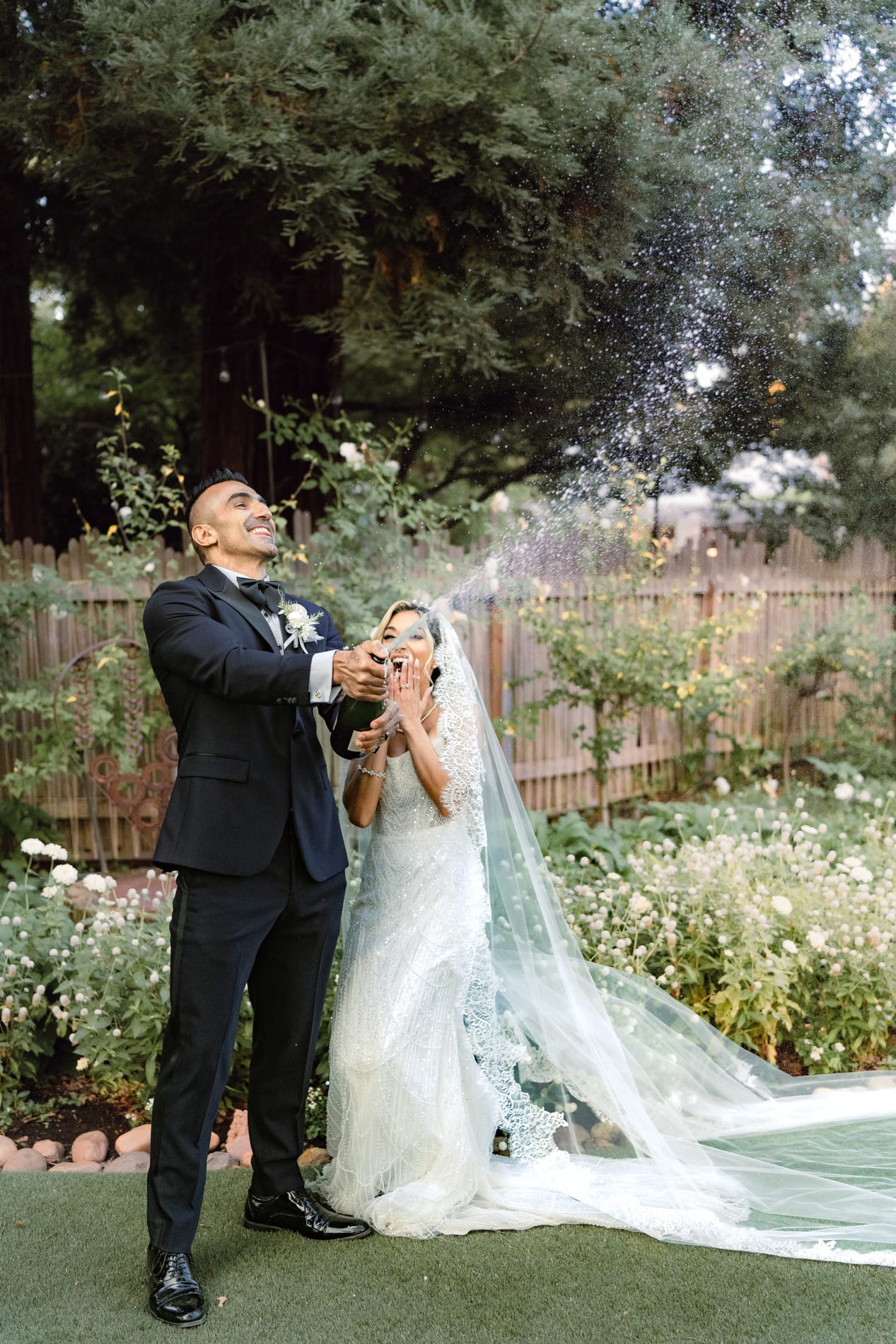Groom spraying champagne over the bride under the redwoods, her veil trailing behind her
