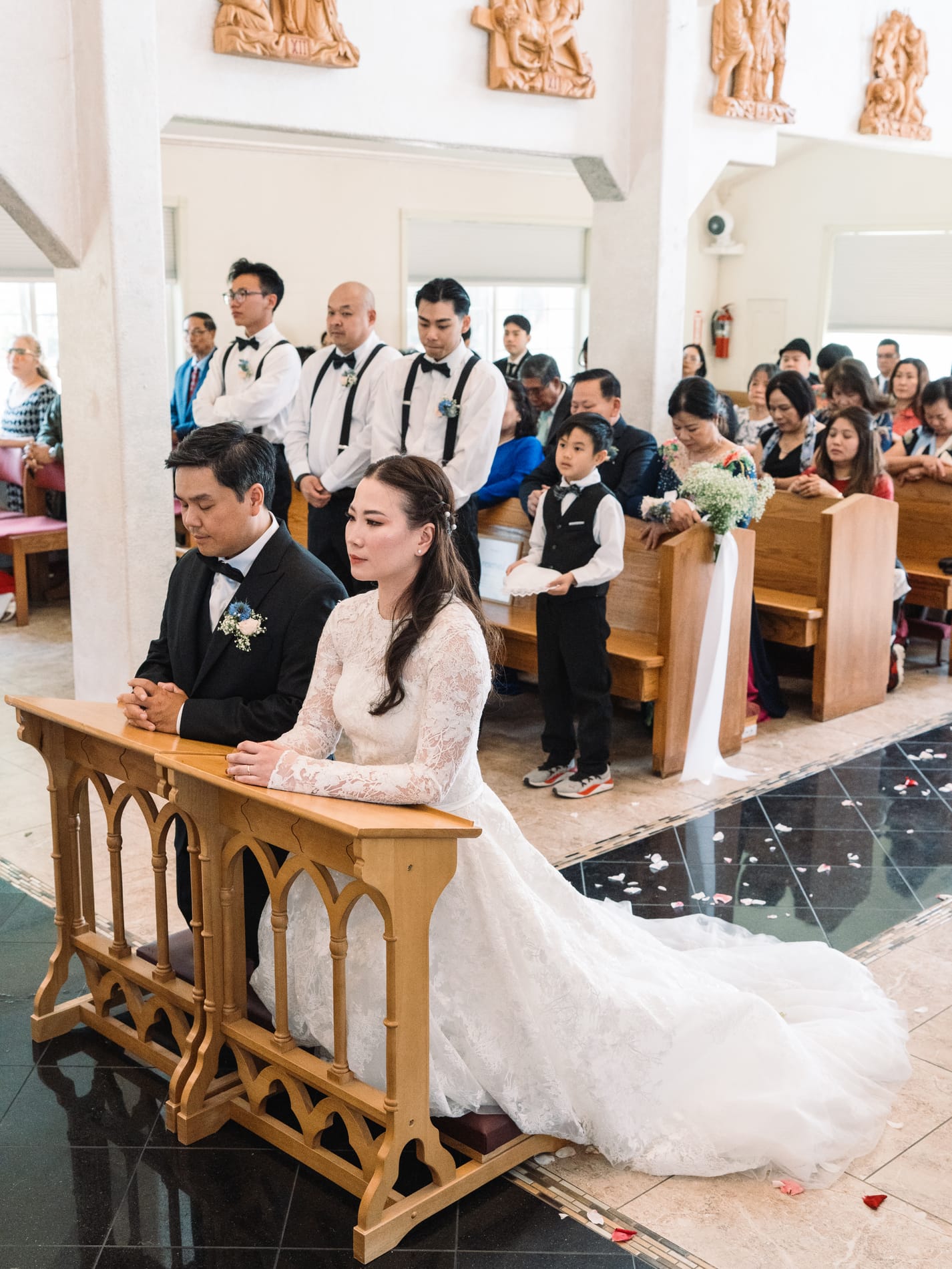 Bride and groom kneeling together during the ceremony, family and wedding party in the pews behind them