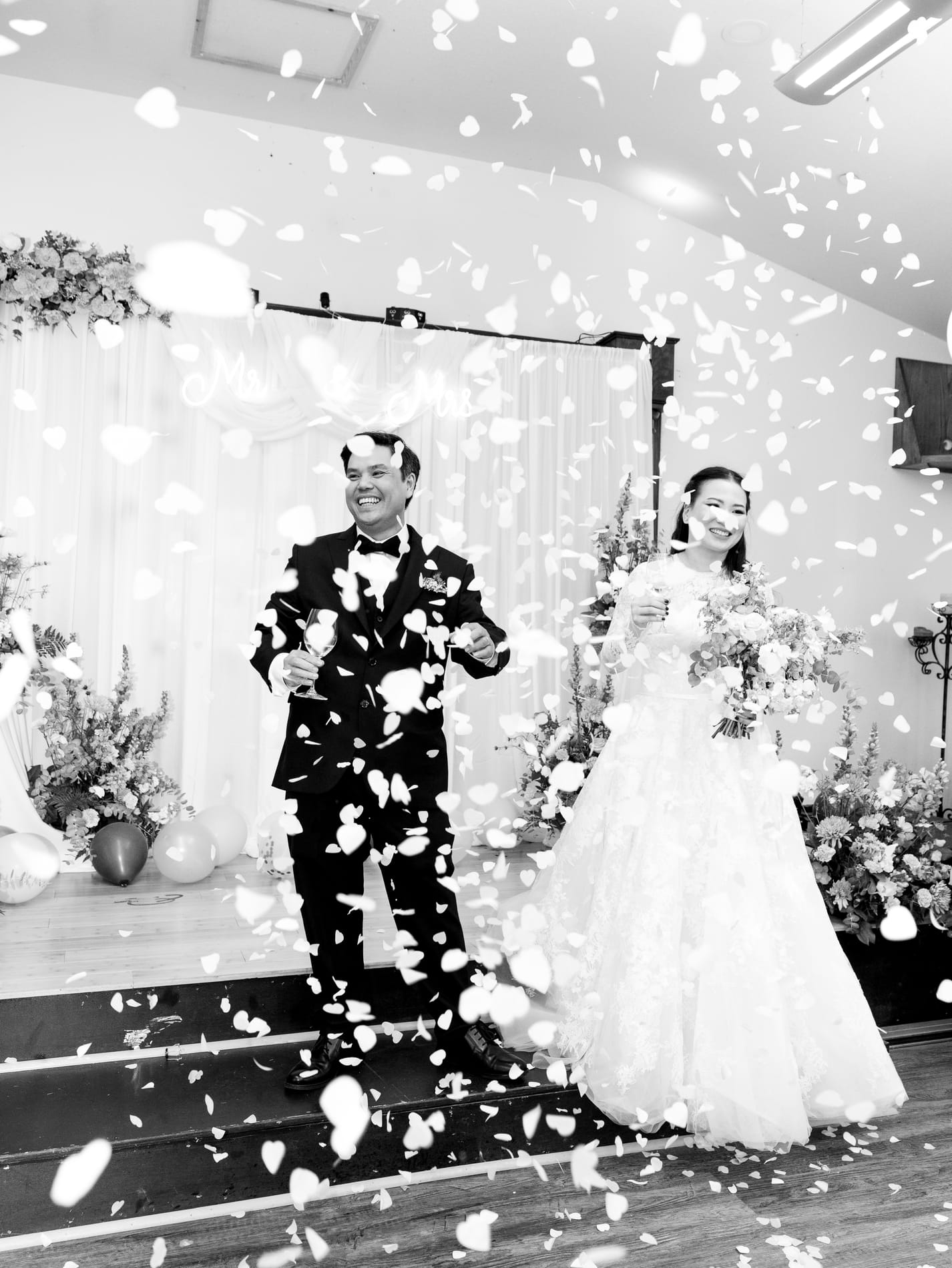 Bride and groom walking out through a shower of petals with champagne in hand, black and white